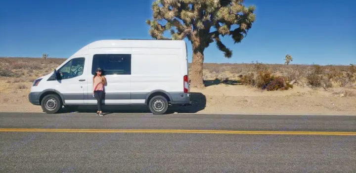 Desert adventure with an RV rental. A woman next to a white camper van on a sunny road, likely Joshua Tree NP.. Ford Transit 2015