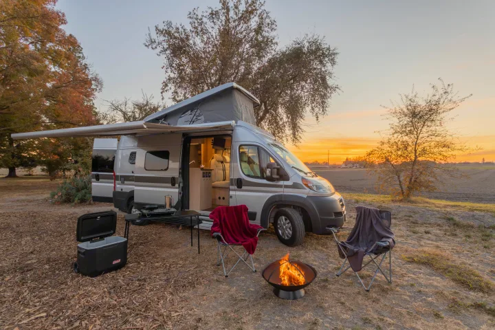 Experience an RV rental: a modern camper van with an open pop-top & awning, camp chairs, cooler, and fire pit at sunset.. Hymer Other 2019