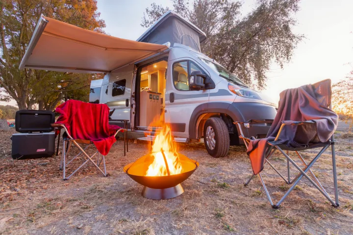 Experience adventure! An RV rental provides a cozy campsite with a lit camper, glowing fire pit, chairs & cooler at dusk.. Hymer Other 2019