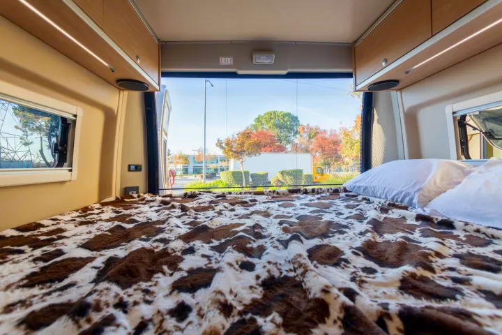 RV rental bed with cow-print blanket, offering a wide rear view of sunny autumn trees and distant buildings.. Hymer Other 2019