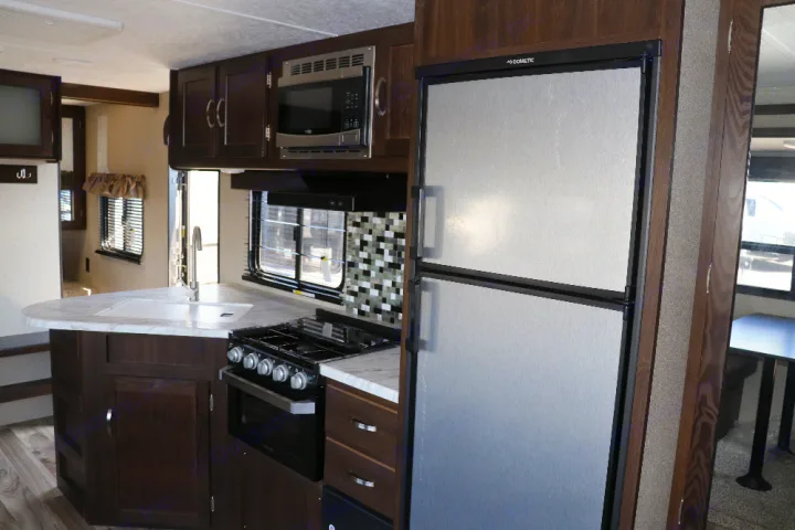 Interior of an RV rental showing a full kitchen with dark cabinets, white counters, stove, microwave, and fridge.. Keystone Springdale 2020