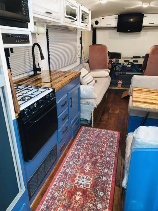 Inside a renovated rv rental: modern kitchen with blue cabinets, wood counter, and stove. A red rug leads to the living and driving areas.. Fleetwood Flair 1997