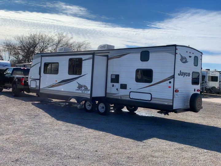 A white Jayco travel trailer with its slide-out extended, connected to a pickup truck on a gravel lot. Ideal for an rv rental adventure.. Jayco Jay Flight 2019
