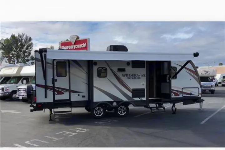 A detailed view of a grey and white travel trailer RV rental, set up with awnings extended, parked in a lot.. Heartland Wilderness 2018