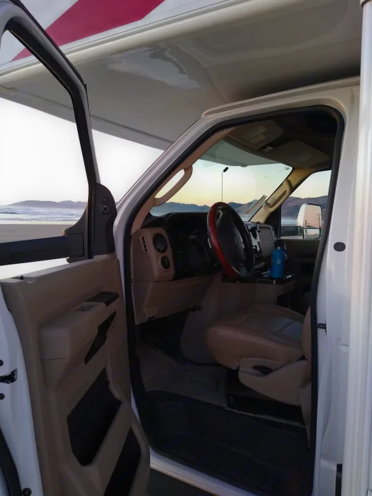 View from an RV rental's cab: driver's seat, steering wheel, & open door revealing a beach with waves & mountains.. Fleetwood Other 2018