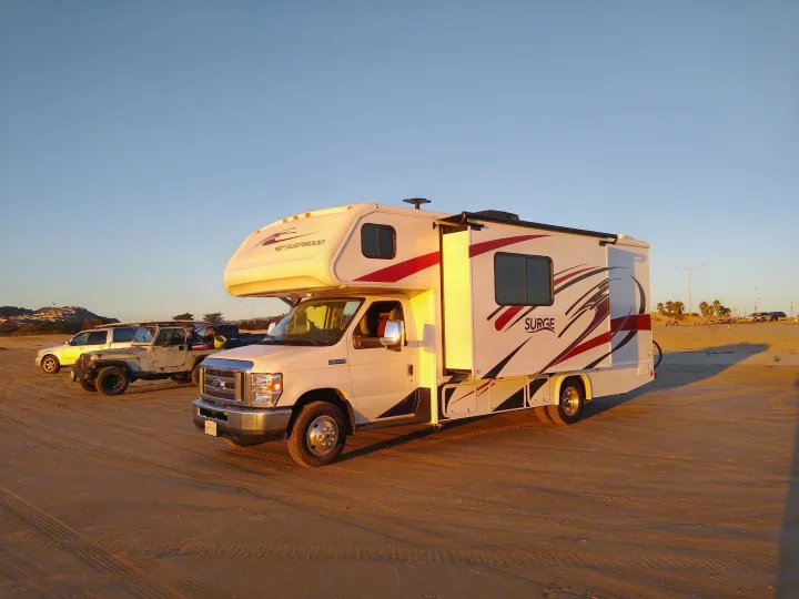 Experience beach camping: A white rv rental parked on a sandy beach at sunset, near other vehicles and distant hills.. Fleetwood Other 2018