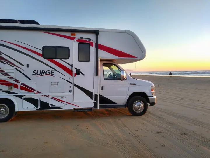 Experience beachside **RV rental** adventures! This white Class C motorhome is parked on a sandy beach at sunset, with a person riding a horse by the ocean.. Fleetwood Other 2018