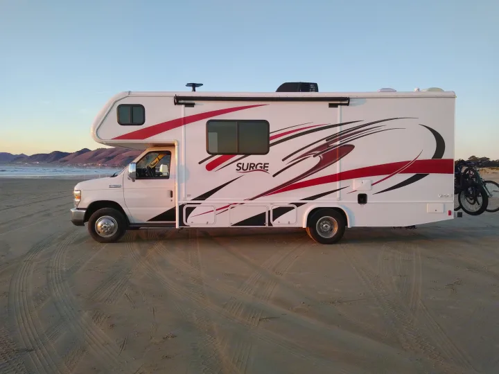 A white SURGE RV rental is parked on a sandy beach, with ocean waves, mountains, and bikes on the rear.. Fleetwood Other 2018