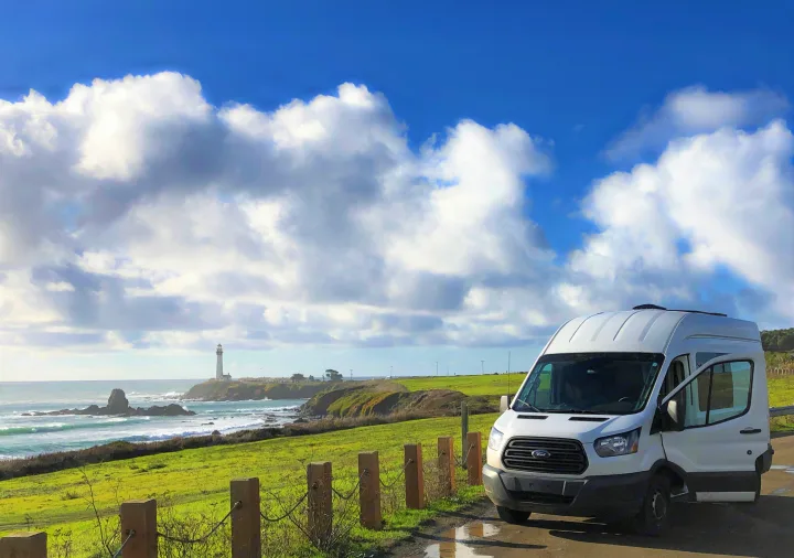White RV rental by the coast with a distant lighthouse, lush green fields, and a blue sky with clouds.. Ford Transit 2015