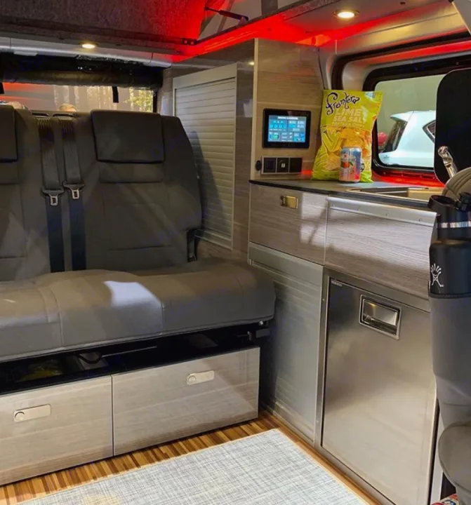 Modern RV rental interior with a compact kitchen, built-in fridge, and comfortable seating that converts to a bed for travel.. Nissan NV200 2019