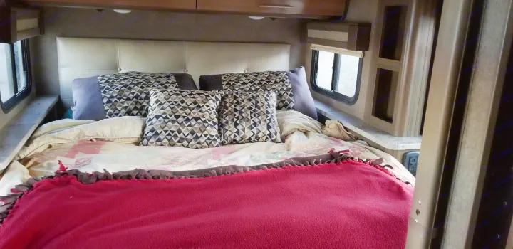 RV rental bedroom: a cozy bed with patterned pillows & red blanket, windows, & built-in storage for comfortable travel.. Thor Motor Coach Gemini 2016