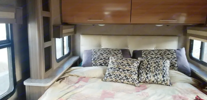RV rental bedroom: a bed with patterned pillows and comforter, light headboard, overhead cabinets, and side windows.. Thor Motor Coach Gemini 2016