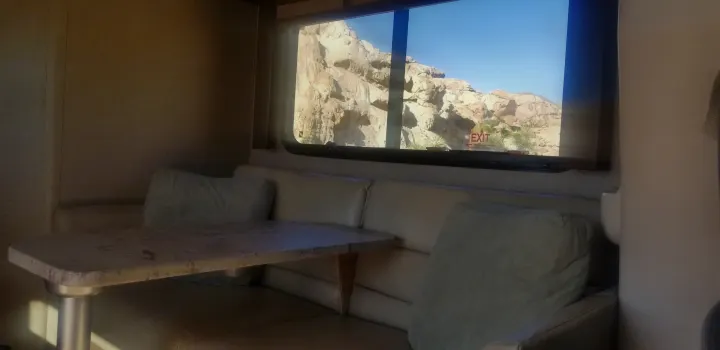 RV rental interior: cozy dinette area with cushions, a table, and a large window showcasing rocky desert scenery. Emergency exit noted.. Thor Motor Coach Gemini 2016