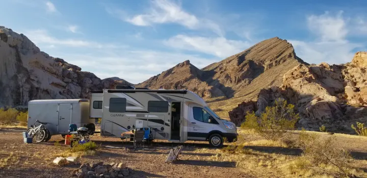 An RV rental and towed trailer are set up for a serene desert campsite with bikes, amidst rugged mountains under a blue sky.. Thor Motor Coach Gemini 2016