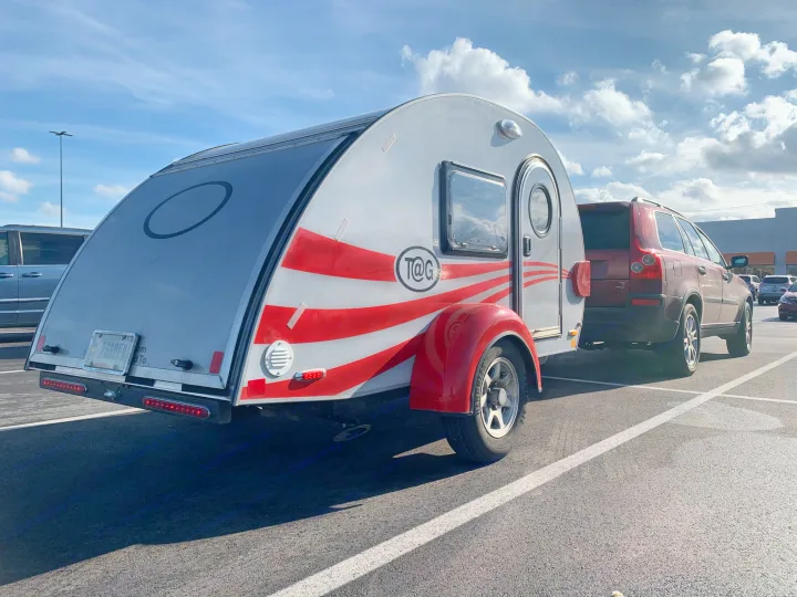 A silver T@G teardrop RV with red stripes, ideal for an rv rental, is hitched to an SUV in a sunny parking lot.. T@G T@G Trailer 2016