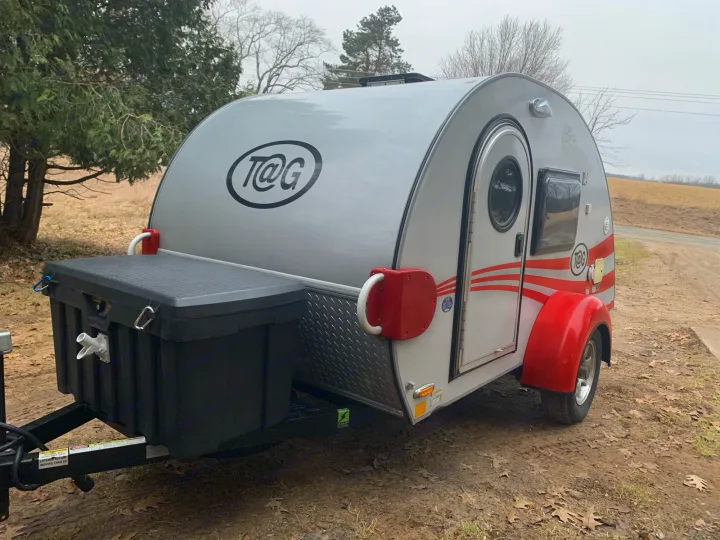Teardrop camper RV rental: Silver and red compact trailer with a front storage box, parked on a rural dirt path.. T@G T@G Trailer 2016