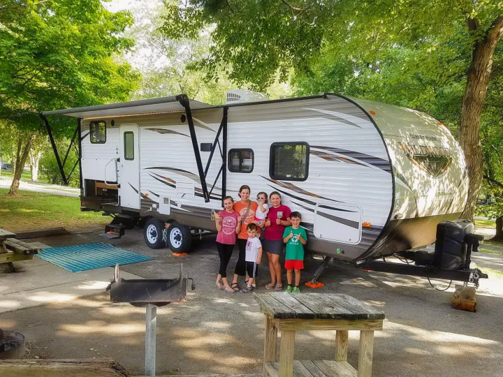 Happy family with their white RV rental at a tree-lined campsite, complete with awning & outdoor setup.. Forest River Wildwood 27DBK 2017