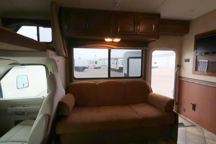 RV rental's inviting interior: comfy sofa, overhead bunk, TV, and large windows revealing other RVs.. Thor Motor Coach Freedom Elite 2013