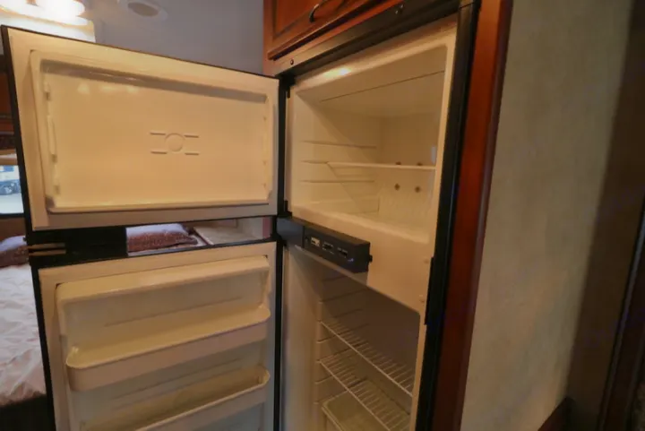 Visually, an open, empty two-door refrigerator within an RV rental. Top is freezer, bottom fridge with shelves.. Thor Motor Coach Freedom Elite 2013