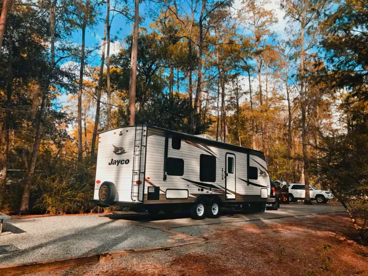 Scenic RV rental: White Jayco travel trailer parked on a gravel campsite amidst tall trees under blue skies.. Jayco Jay Flight SLX 2019