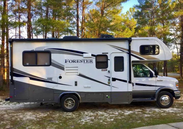 A white and grey Class C RV rental, a large motorhome, parked outdoors on a sandy surface with trees.. Forest River Forester 2018