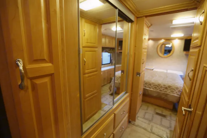 An RV rental interior view. Light wood cabinets and mirrored closet doors lead to a bedroom with a bed and round mirror.. Coachmen Cross Country 2005
