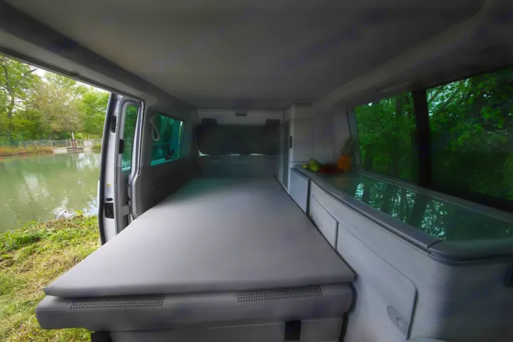 Spacious camper van interior with bed, fruit-laden countertop, and serene lakeside view. Ideal for your next rv rental.. Volkswagen California Coast T6 2021