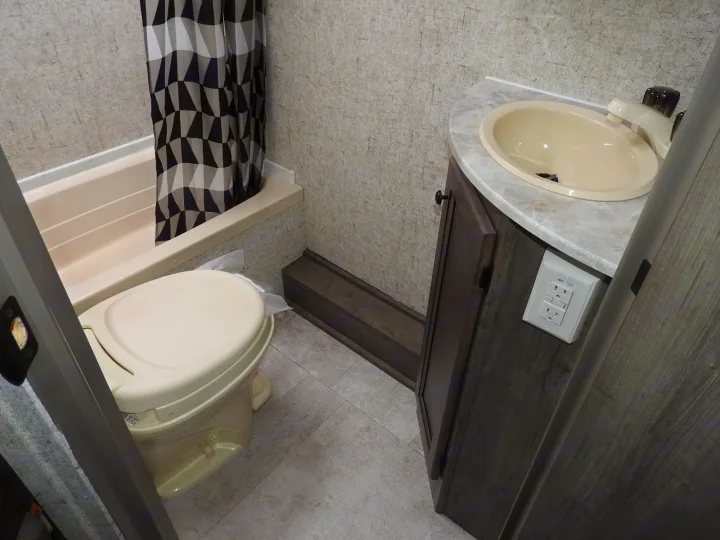 RV rental bathroom features a compact toilet, shower/tub with patterned curtain, and vanity with sink & dark wood cabinet.. Coachmen Apex 208BHS 2023