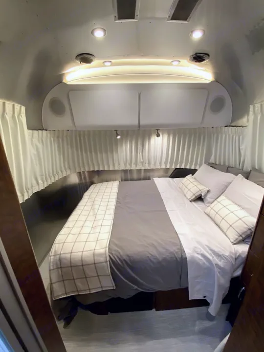Plush RV rental bedroom: full bed with grey bedding, plaid accents, white curtains, and bright overhead lights.. Airstream Globetrotter 2020