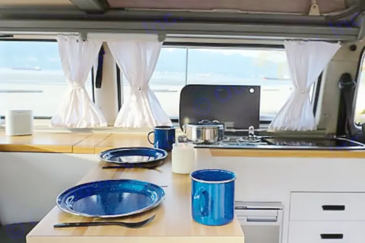 Inside an RV rental: a cozy kitchen and dining area with blue dishes, featuring a window view of the ocean and mountains.. Volkswagen Westfalia 1986