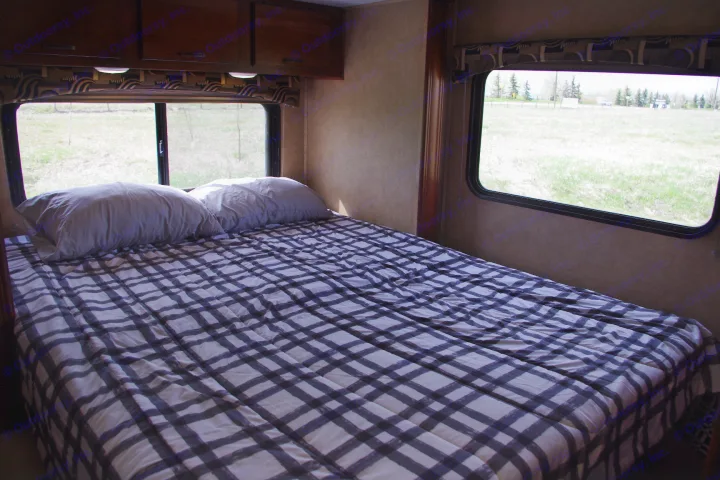 Comfortable RV rental bedroom featuring a plaid bed, pillows, and scenic field views through windows. Ideal for travel.. Forest River Sunseeker 2015