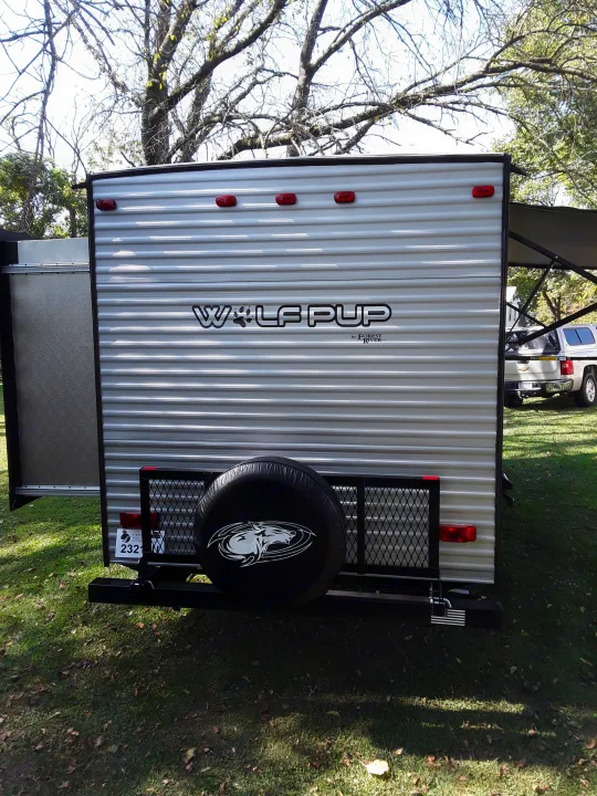 Rear view of a white Wolf Pup travel trailer RV rental, featuring a spare tire with a wolf logo and a cargo rack, parked on grass.. Forest River Wolf Pup 2020