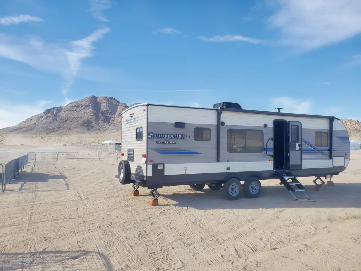 An RV rental trailer is set up on sandy ground near mountains under a bright blue sky with clouds, ready for a desert stay.. K-Z Manufacturing Sportsmen 2019