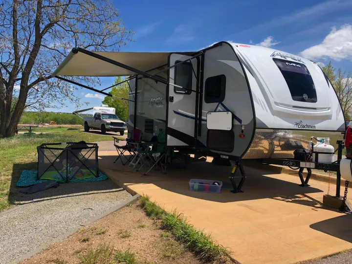 An RV rental is set up at a sunny campsite with an extended awning, outdoor chairs, and a baby's playpen.. Coachmen Apex Nano 2019