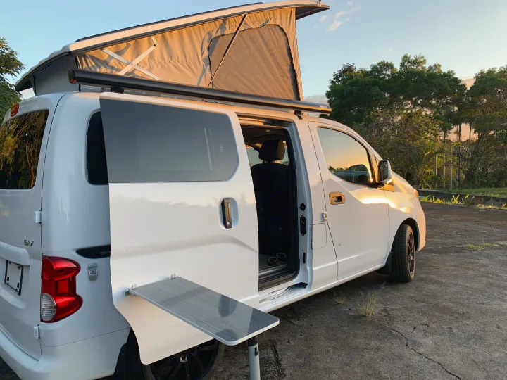 White pop-top camper van, with side door open and fold-out table, parked at sunset. Perfect RV rental for adventure.. Nissan NV200 2019