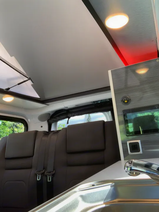 RV rental interior: Overhead cabinet with screen, power outlet, lighting. Comfy seating with seatbelts & a sink are also visible.. Nissan NV200 2019