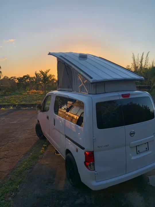 White Nissan NV200 camper van with pop-top extended at sunset, ready for an RV rental road trip in a tropical setting.. Nissan NV200 2019