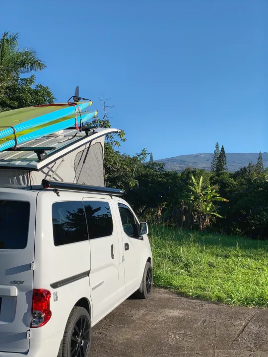 White pop-top RV rental campervan with paddleboards on its roof, parked amidst lush greenery with mountains in the background.. Nissan NV200 2019