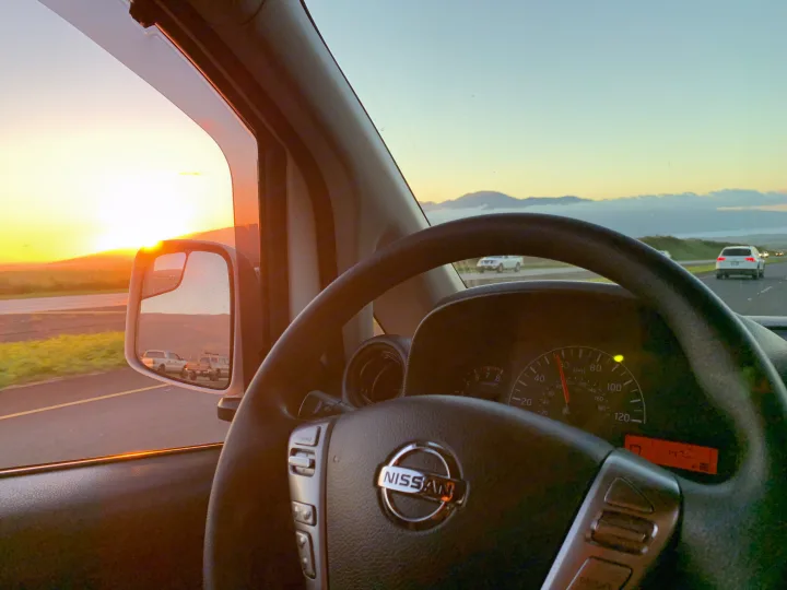 Golden sunset view from a Nissan car. Dashboard and side mirror show road & mountains, evoking a scenic RV rental road trip.. Nissan NV200 2019