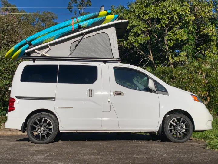 White pop-top camper van with two paddleboards on top, ready for adventure. Explore the outdoors with this RV rental!. Nissan NV200 2019