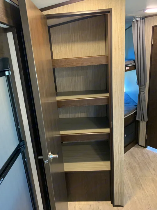 Explore an RV rental's interior: an open pantry with multiple shelves, a partial view of a refrigerator, and bunk beds with a privacy curtain.. Cruiser Rv Corp Shadow Cruiser 2019