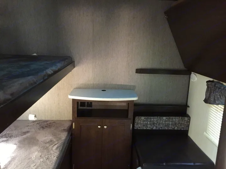 Compact RV rental interior featuring dark bunk beds, a central storage unit, and a black dinette with a patterned cushion.. Heartland Trail Runner 2020