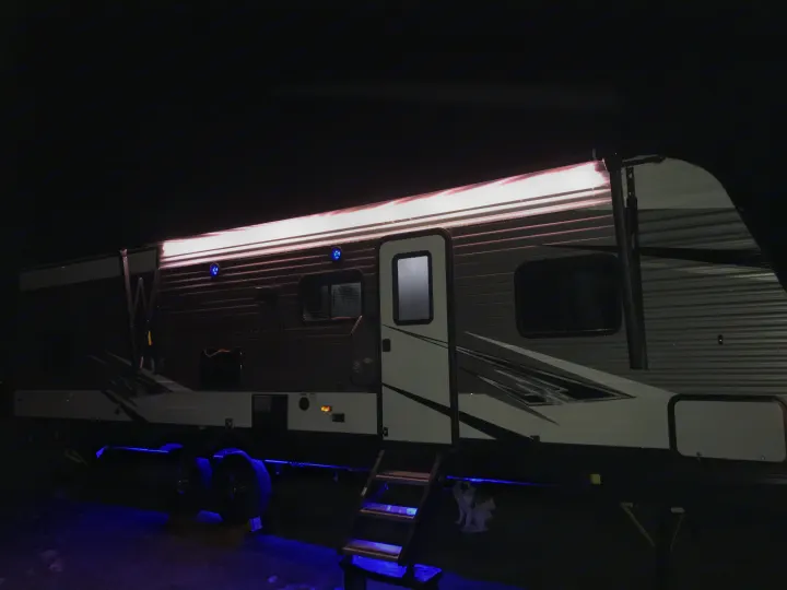 RV rental glowing at night with bright awning lights & blue ground effects, perfect for cozy camping. Dog visible near steps.. Heartland Trail Runner 2020