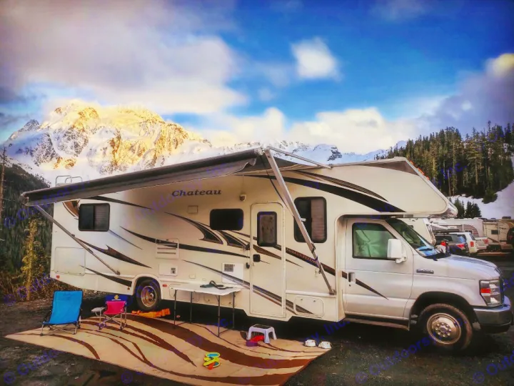 An RV rental is parked with an extended awning and outdoor setup at the base of stunning sunlit snow-capped mountains.. Ford Fourwinds 2012