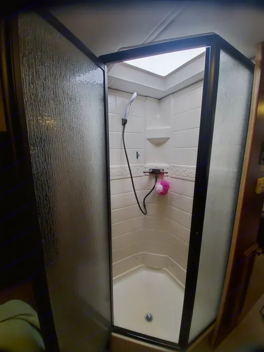 Compact RV rental shower with clear textured door & skylight. White interior, handheld shower, and loofah visible.. Ford Fourwinds 2012