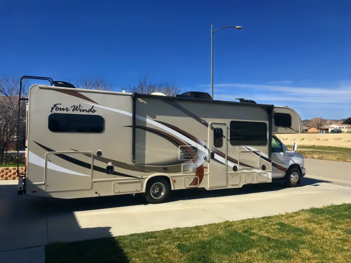 A light-colored Class C Four Winds RV, ideal for an RV rental, is parked on a paved driveway under a clear blue sky, ready for adventure.. Thor Motor Coach Four Winds 2017
