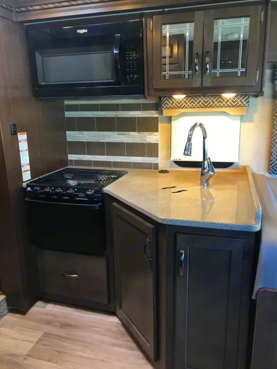 RV rental kitchen: dark wood cabinets, light countertop, black microwave, gas stove, and sink. Perfect for your next adventure.. Thor Motor Coach Four Winds 2017