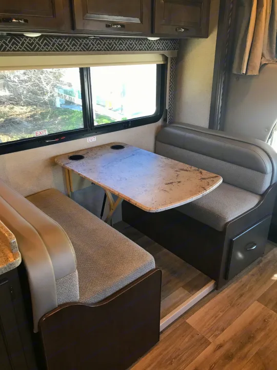 View inside an RV rental showing a light-colored dinette with opposing benches, a table, and window with "EXIT" sign.. Thor Motor Coach Four Winds 2017