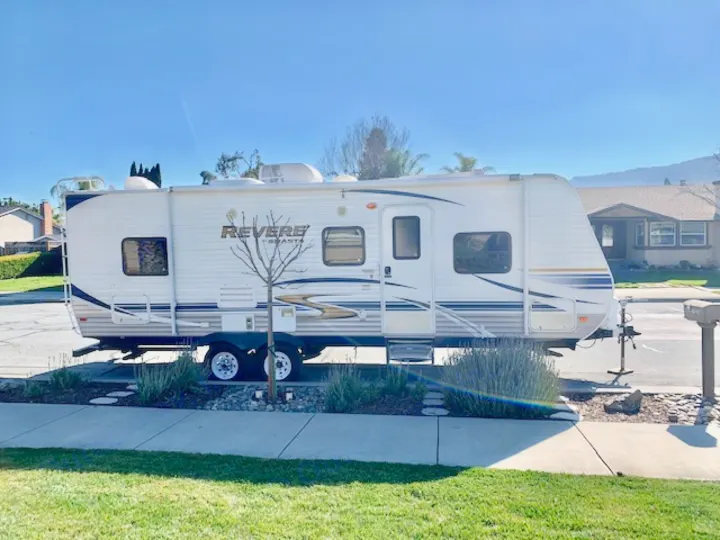 A white Revere Shasta travel trailer, an RV rental, parked on a sunny residential street. Blue sky above houses & greenery.. Shasta Revere 2012