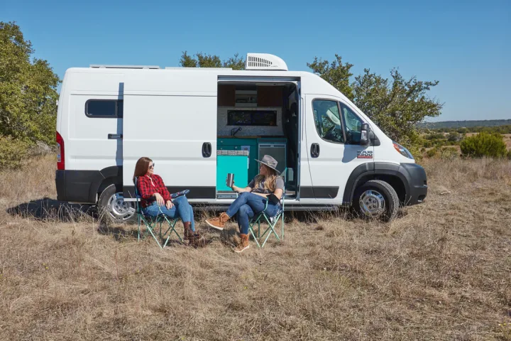 Relaxing RV rental getaway: Two women sit in camping chairs beside their white camper van in a sunny field. #rvrental #vanlife. Dodge 2500 Promaster 2019